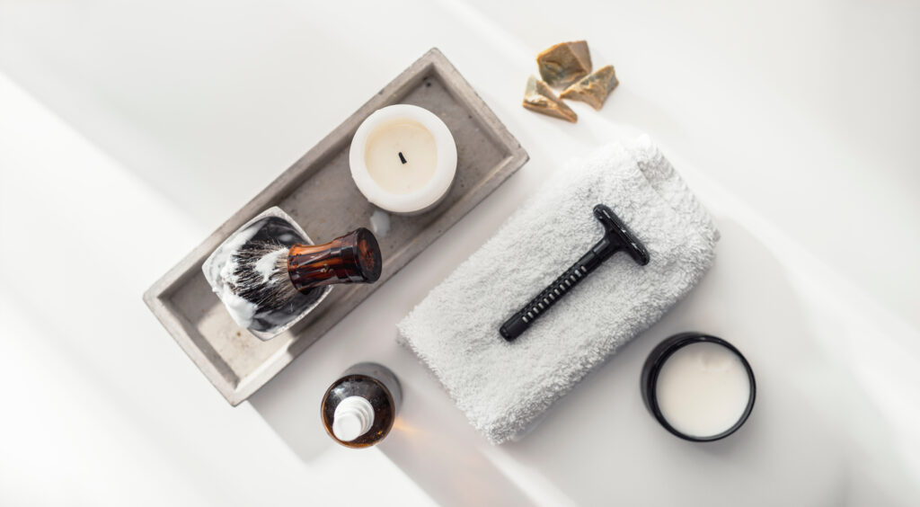 Personal grooming essentials arranged on a light surface, including a razor, towel, shaving brush, and candle