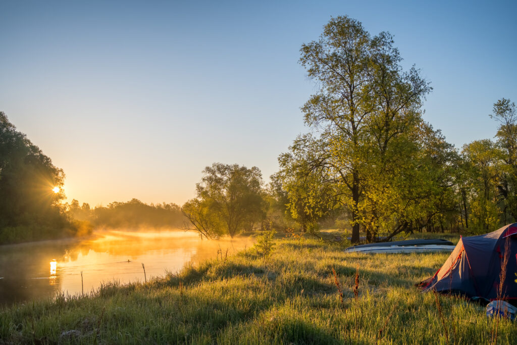 Tent set up beside a river at sunrise with trees and mist along the shoreline